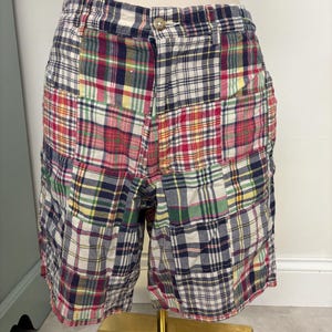 May include: Patchwork shorts featuring a multi-colored plaid pattern in shades of red, navy blue, green, yellow, and white. The shorts have a button closure at the waist, a zip fly, and a classic knee-length cut with a structured waistband.