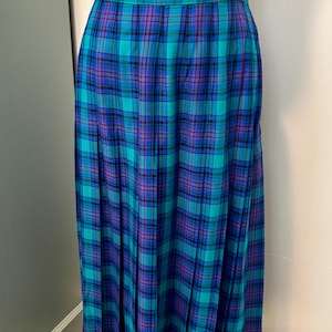 May include: A floor-length pleated skirt featuring a plaid pattern in shades of teal, royal blue, and purple. The skirt has a fitted waistband and a straight silhouette that falls to the ankles, showcasing a consistent tartan-inspired grid design.