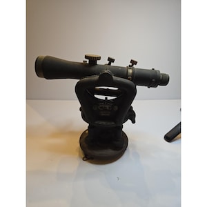 May include: A vintage black metal surveying level instrument featuring a horizontal telescope with multiple adjustment knobs, a central support frame, and a circular base with leveling screws. The device displays visible markings and dials.