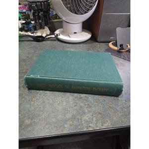 May include: A green hardcover book titled Basic Physics of Radiation Therapy with gold lettering on the spine. The book has a textured fabric cover and is positioned horizontally, showing the front cover and the spine with the printed title.