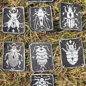 May include: A collection of eight rectangular black fabric patches, each featuring a distinct white screen-printed insect design. The insects include various beetles, a moth, and a pill bug, all rendered in a high-contrast, graphic style.