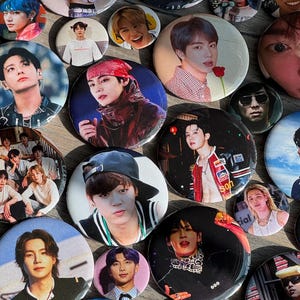 May include: A collection of circular pin-back buttons featuring various photographic portraits of the members of the K-pop group BTS. The buttons display individual close-ups and group shots of the performers in diverse outfits and settings.