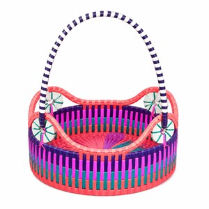 May include: A round woven basket featuring a vibrant multi-colored pattern of pink, purple, and teal vertical strips. The basket includes a tall, arched handle with navy blue and white stripes and four circular white accents with green stitching.