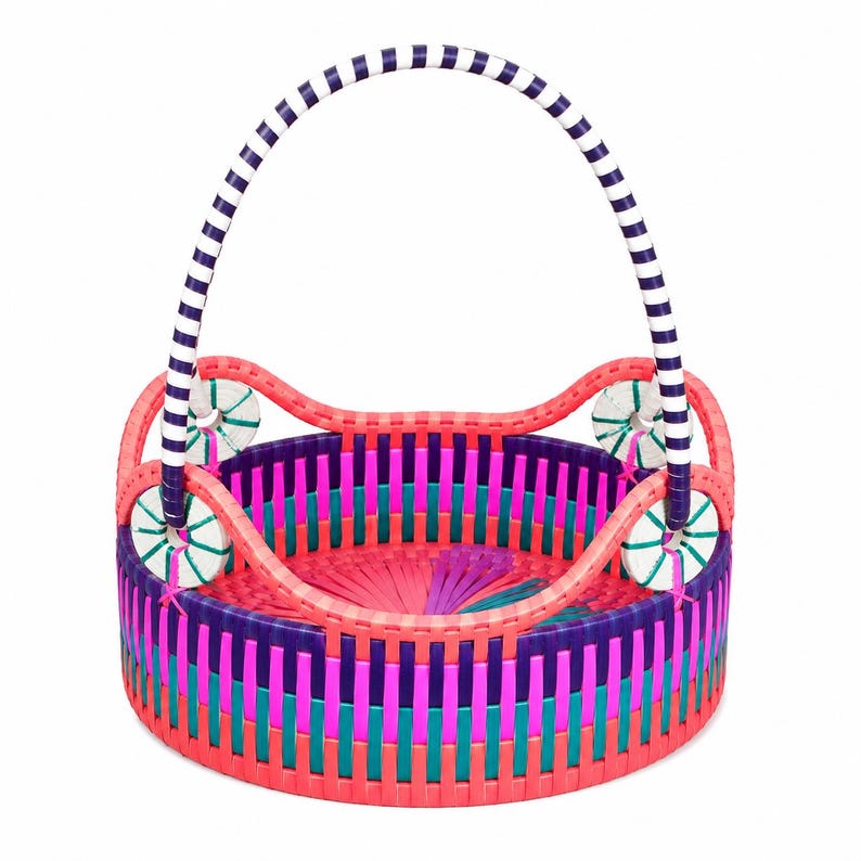 May include: A round woven basket featuring a vibrant multi-colored pattern of pink, purple, and teal vertical strips. The basket includes a tall, arched handle with navy blue and white stripes and four circular white accents with green stitching.