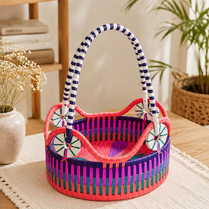 May include: A round woven basket featuring a vibrant multi-colored pattern of pink, purple, and teal vertical strips. It has a high, arched handle with blue and white stripes and four circular decorative accents where the handle meets the rim.