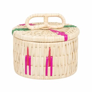 May include: A round woven basket with a lid featuring a central handle. The basket is constructed from light beige natural fibers and accented with vertical stripes of bright pink and green woven into the sides and the top of the lid.