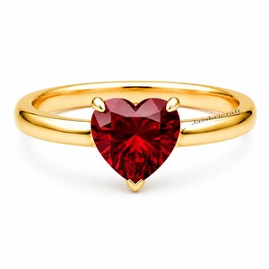 Op de afbeelding: Een gouden ring met een centrale hartvormige rode edelsteen vastgehouden door drie pootjes. De ringband is van glad en gepolijst goud. De tekst Jaishricraft is zichtbaar aan de rechterkant van de band. Het ontwerp is gecentreerd op een witte achtergrond.
