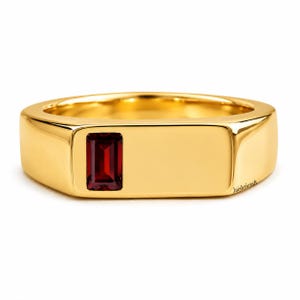 May include: A polished gold signet ring featuring a single rectangular, deep red garnet gemstone set flush into the flat top surface. The band has a smooth, wide, and rounded profile. The text Jaishricraft is printed on the side of the ring.