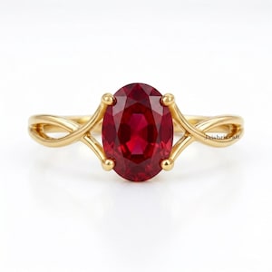 May include: A gold ring featuring a large, oval-cut, deep red gemstone at the center. The band has a polished gold finish and a split, twisted design that connects to the central setting. The text Jaishricraft is visible on the band.