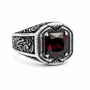 May include: A silver ring featuring a square-cut dark red gemstone set in a bezel with four prongs. The band is decorated with intricate black-oxidized scrollwork engravings and includes the text Jaishricraft printed on the side surface.