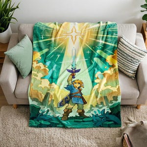 May include: A soft fleece blanket featuring a vibrant illustration of a blonde character in a blue tunic holding a glowing sword aloft. The background displays a bright starburst, stylised clouds, and green landscape elements in shades of yellow and teal.