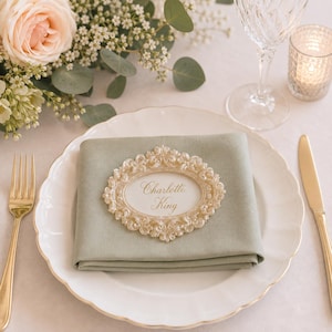 May include: A sage green cloth napkin folded on a white scalloped plate, featuring an oval cream-colored ornate frame with the name Charlotte King printed in gold script. A gold fork and knife are placed on either side of the plate.