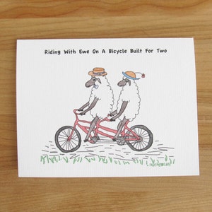May include: Two white sheep wearing hats ride a red tandem bicycle with a green grass background. The text "Riding With Ewe On A Bicycle Built For Two" is at the top of the image.