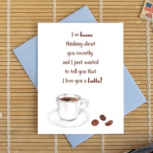 May include: A greeting card with the text "I've bean thinking about you recently, and I just wanted to tell you that I love you a latte!" features a coffee cup illustration. The card is on a blue envelope, with stamps and a pen.