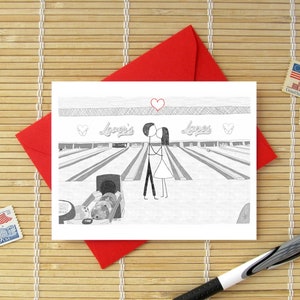May include: A white greeting card featuring a black and white line drawing of two stick figures kissing at a bowling alley. The text "Lover's Lanes" is written above the bowling lanes, with a red heart drawn above the figures and two smaller hearts on the sides.
