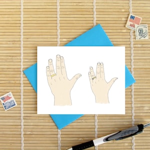 May include: Greeting card with a hand-drawn illustration of two hands in the Vulcan salute, each wearing a gold ring. The card is on a blue envelope, with stamps and a pen nearby, set against a bamboo mat.