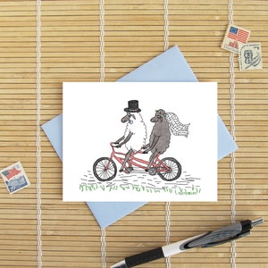 May include: A white sheep wearing a top hat and a black sheep wearing a flower crown ride a red tandem bicycle. The sheep are smiling and the bicycle has a basket on the back. The text "till death do us part" is written below the image.