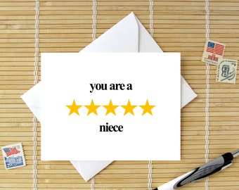 May include: A white greeting card with the text "you are a niece" and five yellow star ratings. The card is inside a white envelope, with a pen and postage stamps nearby. The background is a bamboo mat.