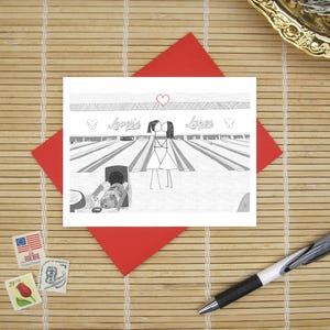 May include: A white greeting card featuring a black and white line drawing of two stick figures kissing at a bowling alley. The text "Lover's Lanes" is written above the figures, with a small red heart centered above the text.