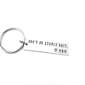 May include: A silver metal keychain with a rectangular tag. The tag has the phrase "DON'T DO STUPID SHIT. ♡ MOM" in black, sans-serif font. The keychain ring is circular.