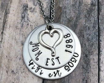 May include: Silver necklace with a heart charm and two layered discs. The top disc has the text "MOM EST" and the year "1988". The bottom disc has the text "Gigi est" and the year "2020".