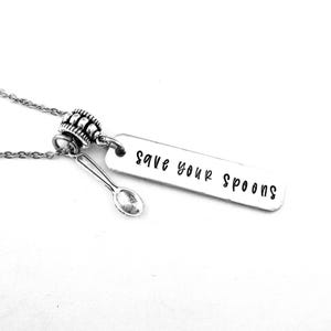 May include: A silver-toned necklace featuring a thin chain, a textured cylindrical bead, a small spoon charm, and a rectangular metal pendant stamped with the black text "save your spoons" in a simple, lowercase sans-serif font.