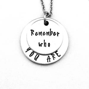 May include: A silver-colored pendant necklace with two circular charms. The top charm has the words "Remember who" and the bottom charm says "YOU ARE" in black handwritten font. The necklace chain is also silver.