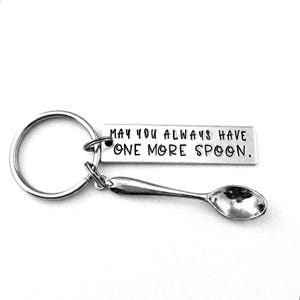 May include: A silver keychain with a spoon charm and a rectangular tag. The tag reads "MAY YOU ALWAYS HAVE ONE MORE SPOON." The keychain is on a white background.