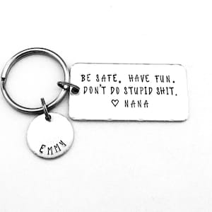 May include: A silver-colored keychain with a rectangular tag and a small round tag. The rectangular tag has the text "BE SAFE. HAVE FUN. DON'T DO STUPID SHIT. ♡ NANA". The round tag has the name "EMMY".