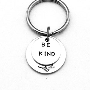 May include: Silver keychain with a double-sided charm. The top side reads "BE KIND" and the bottom side has a small floral design.