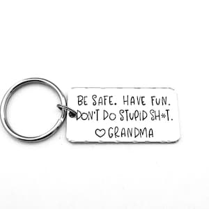 May include: A silver-colored keychain with a rectangular tag. The tag has the text "BE SAFE. HAVE FUN. DON'T DO STUPID SH*T. ❤️ GRANDMA" in black lettering. The keychain ring is attached to the tag.