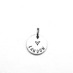May include: A small, round, silver-colored metal tag with a keyring at the top. The tag has the name "LANDON" in a handwritten font and a small heart symbol. The background is plain white.