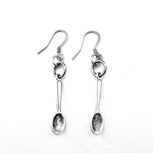 May include: A pair of silver-toned metal earrings featuring a miniature spoon charm dangling from a circular jump ring. Each earring is attached to a curved ear wire hook, showcasing a textured, antique-style finish on the spoon bowl.