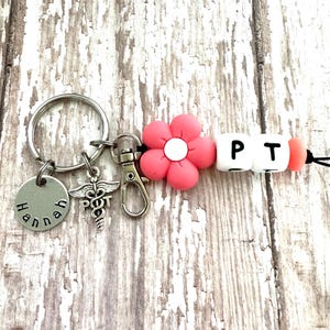 May include: A pink flower charm with a white center, a silver charm with a medical symbol, a silver charm with the name "Hannah" engraved on it, and three white letter beads spelling "PT" on a silver key chain.