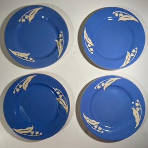 Könnte beinhalten: Ein Set aus vier runden Keramiktellern in leuchtendem Blau. Jeder Teller ist mit einem cremefarbenen Blumenmuster aus Maiglöckchenzweigen verziert, die entlang der Ränder angeordnet sind und ein einheitliches Design für alle vier Stücke bilden.