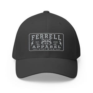 May include: A black baseball cap featuring a white rectangular patch on the front. The patch displays a raccoon illustration, lightning bolts, and the text FERRELL APPAREL EST. 2026 STAY WILD in a distressed, vintage-style font and graphic design.