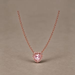 May include: A rose gold necklace featuring a heart-shaped pendant with a central light pink gemstone surrounded by a halo of small clear stones. The pendant is suspended from a delicate rose gold chain with a classic link design.