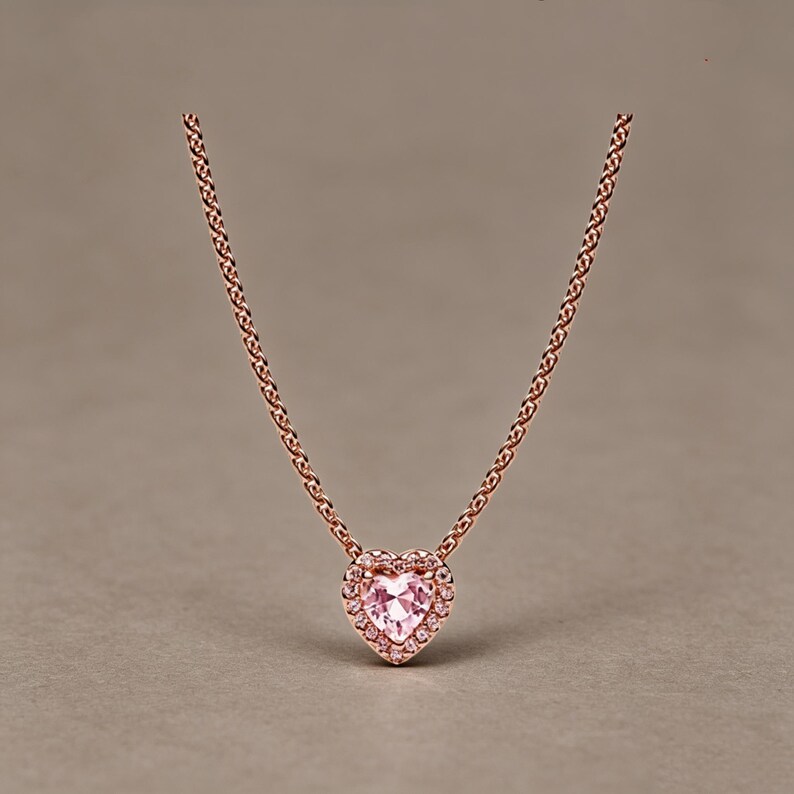 May include: A rose gold necklace featuring a heart-shaped pendant with a central light pink gemstone surrounded by a halo of small clear stones. The pendant is suspended from a delicate rose gold chain with a classic link design.