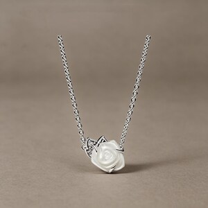 May include: A silver chain necklace featuring a central pendant shaped like a white rose with a glossy finish. The rose is accented by a small silver leaf detail embedded with tiny clear stones, creating a delicate and refined floral jewellery piece.