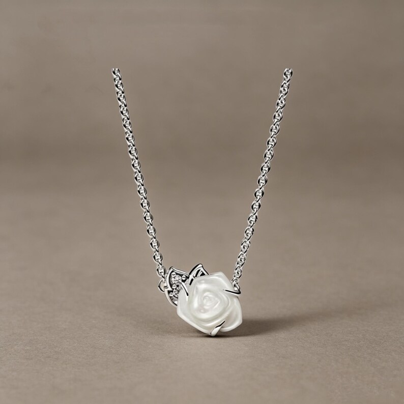 May include: A silver chain necklace featuring a central pendant shaped like a white rose with a glossy finish. The rose is accented by a small silver leaf detail embedded with tiny clear stones, creating a delicate and refined floral jewellery piece.