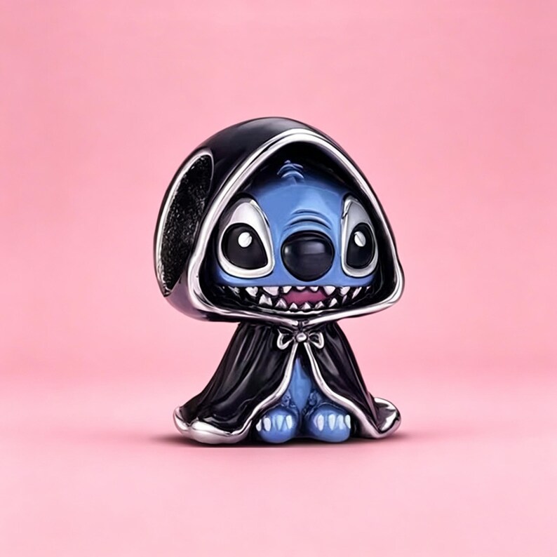 May include: A metal charm bead featuring a blue character with large black eyes and a wide grin, wearing a black hooded cloak with silver trim. The character has pointed ears and a small nose, with the cloak draped over its shoulders and tied.