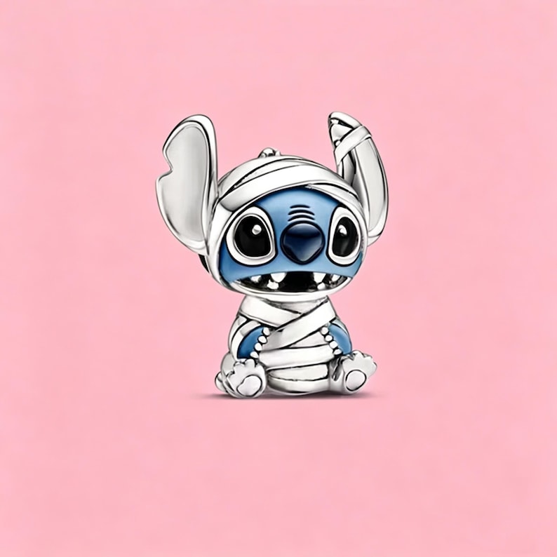 May include: A silver-toned metal charm shaped like the character Stitch, depicted with blue skin and large black eyes, wrapped in white bandages across the head and body. The charm features a glossy finish with detailed engraved lines.