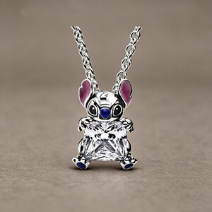 May include: A silver-toned pendant necklace featuring a character with large ears, pink inner ear accents, and a dark blue nose, holding a large, square-cut, clear faceted gemstone. The pendant hangs from a delicate silver-toned metal chain link.