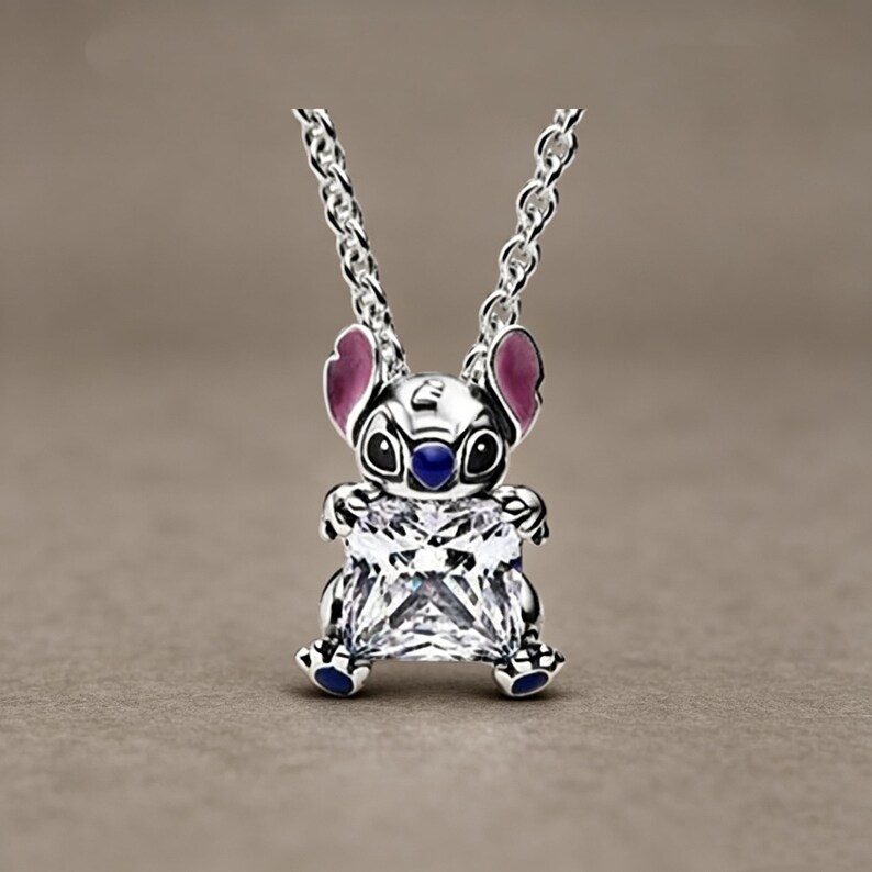 May include: A silver-toned pendant necklace featuring a character with large ears, pink inner ear accents, and a dark blue nose, holding a large, square-cut, clear faceted gemstone. The pendant hangs from a delicate silver-toned metal chain link.