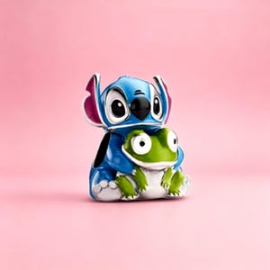 May include: A metal charm featuring a blue character with large ears and pink inner accents, embracing a small green frog with white underbelly details. The charm has a glossy finish and is designed with a hole on the side for attachment to jewelry.