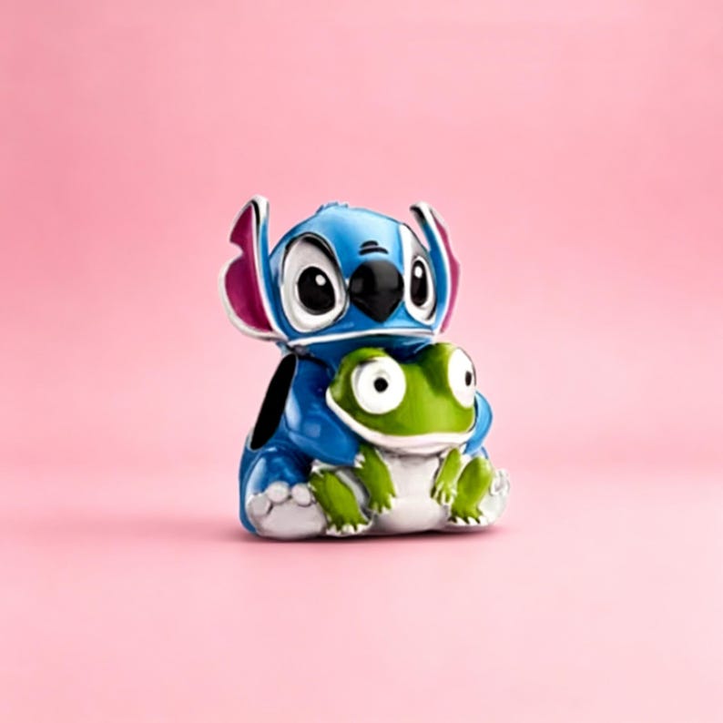 May include: A metal charm featuring a blue character with large ears and pink inner accents, embracing a small green frog with white underbelly details. The charm has a glossy finish and is designed with a hole on the side for attachment to jewelry.