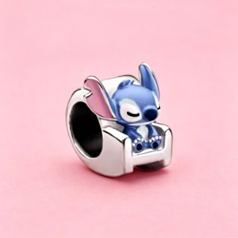 May include: A silver charm bead featuring a blue and pink character sitting in a silver armchair. The character has large ears, a dark blue nose, and closed eyes. The charm has a hollow center for a bracelet, with a polished metallic finish.