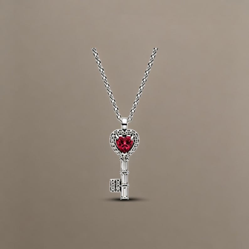 May include: A silver-toned necklace featuring a key-shaped pendant. The head of the key is a heart shape set with a vibrant red gemstone, surrounded by small clear stones. The shaft of the key is accented with baguette-cut clear stones.