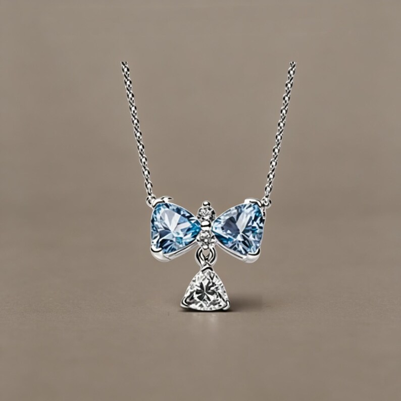May include: A silver necklace featuring a bow-shaped pendant set with two light blue triangular gemstones and a small clear stone in the centre. A single clear triangular gemstone hangs below the bow, suspended from a delicate silver chain.