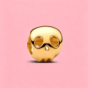 May include: A polished gold-tone metal bead shaped like a stylized skull. The bead features smooth, rounded contours with two large, hollowed-out eye sockets and a small, triangular nasal cavity, designed with a central hole for threading.